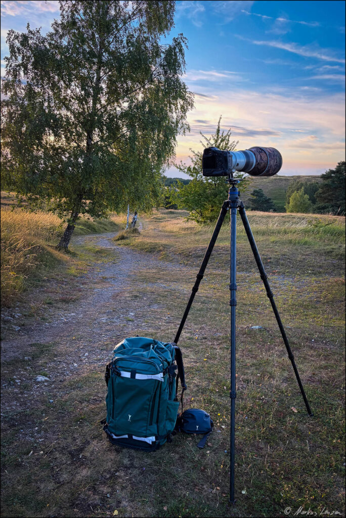 Kamera mit Teleobjektiv auf einem Carbon-Stativ neben dem NYA-EVO Fjord 80L Rucksack in einer Hügellandschaft.